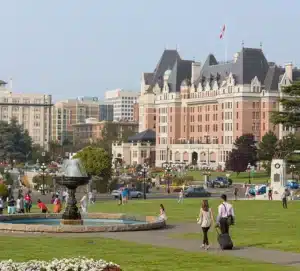 People walk and relax on a grassy area with a fountain in front of a large historic hotel building, with city buildings in the background.