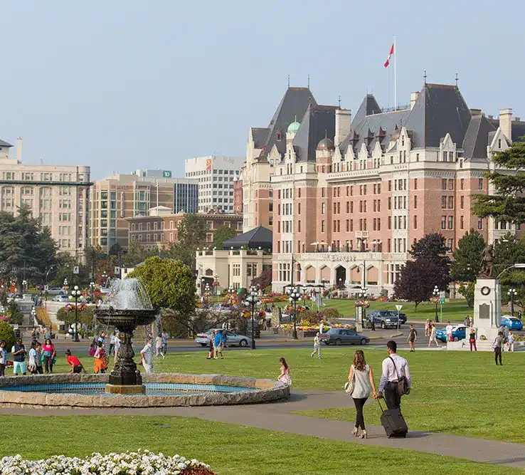 People walk and relax on a grassy area with a fountain in front of a large historic hotel building, with city buildings in the background.