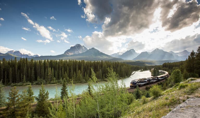 A train travels along tracks beside a winding river, surrounded by dense forest and mountains under a partly cloudy sky.