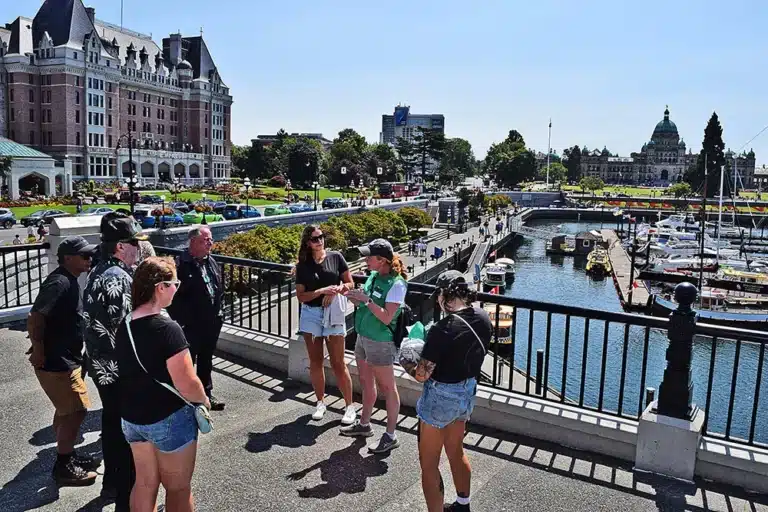A group of people stands on a waterfront path overlooking a marina, with historic buildings and parked cars visible in the background on a sunny day.