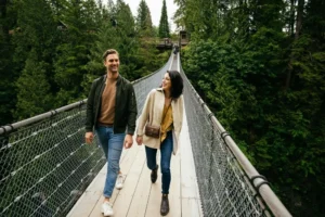 Two people walk and smile on a suspension bridge surrounded by dense green forest.
