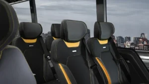 Four black and yellow high-back seats with headrests and seatbelts are arranged in a row inside a vehicle, with city buildings visible through large windows.