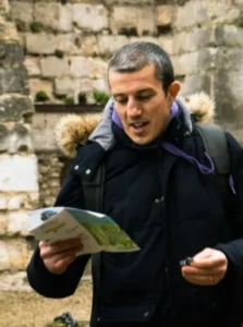 A person wearing a dark jacket with a fur-lined hood reads a brochure while holding a small object, standing in front of a stone wall.