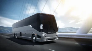 A large, modern Prevost coach bus drives on a bridge with mountains and a bright sky in the background.
