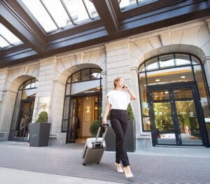 Woman in business attire walks with a rolling suitcase outside a modern building, talking on her phone.