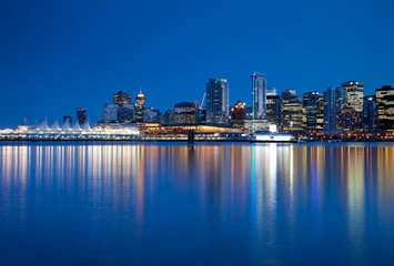 City skyline at night with illuminated buildings reflecting on calm water under a clear, dark blue sky.