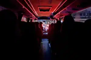 Interior of a dimly lit bus at night with passengers seated, illuminated by red lights and city traffic visible through the front windshield.
