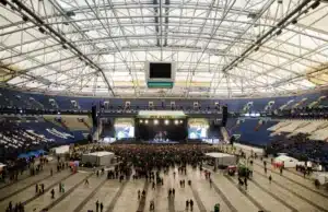 A large indoor stadium with a crowd gathered in front of a stage for a concert, surrounded by rows of mostly empty blue seats.
