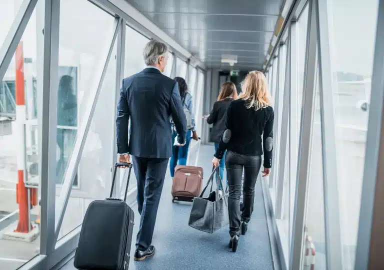 People walking down an airport jet bridge, carrying luggage and bags, heading towards an airplane.