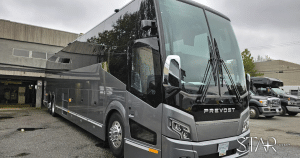 A large gray Prevost tour bus is parked on a paved lot near industrial buildings under an overcast sky.