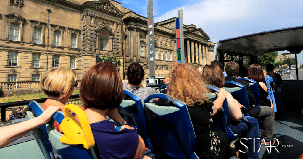 A group of people sit on the upper deck of an open-top tour bus passing historic stone buildings on a sunny day.