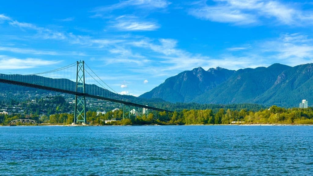 A suspension bridge spans across blue water with green trees and mountains in the background under a clear blue sky, making it a perfect spot for Vancouver sightseeing during your bus tour.