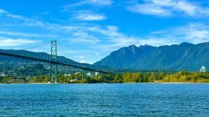 A suspension bridge spans across blue water with green trees and mountains in the background under a clear blue sky, making it a perfect spot for Vancouver sightseeing during your bus tour.