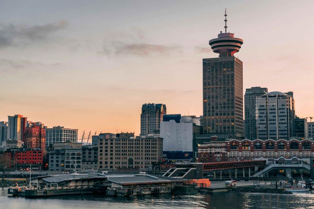 Downtown city skyline at sunset featuring the Vancouver Lookout tower, with waterfront buildings and calm water in the foreground—a perfect view to enjoy on a Vancouver bus tour.