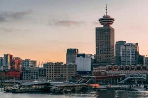 Downtown city skyline at sunset featuring the Vancouver Lookout tower, with waterfront buildings and calm water in the foreground—a perfect view to enjoy on a Vancouver bus tour.