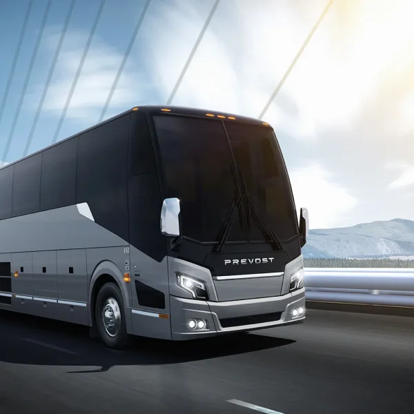 A modern Prevost coach bus drives on a bridge with mountains and water visible in the background under a partly cloudy sky.
